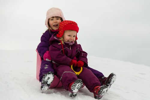 Two children in winter clothing joyfully sledding down a snowy hill, smiling and enjoying the winter fun.