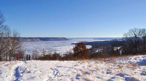 A snowy landscape with a frozen lake and distant hills under a clear blue sky, featuring hikers in the foreground.