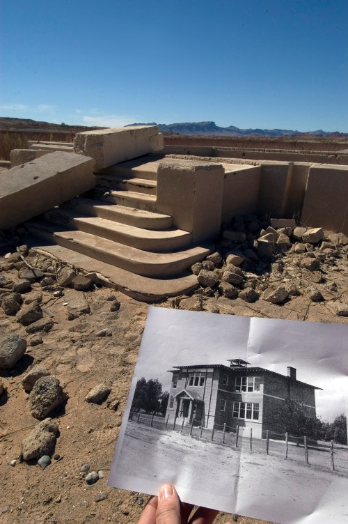 A person holds a black-and-white photo of a building in front of its ruins, with steps and rocky terrain visible.