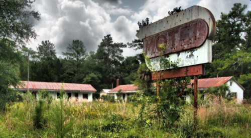 An abandoned motel with a weathered sign, surrounded by overgrown grass and trees under a cloudy sky.