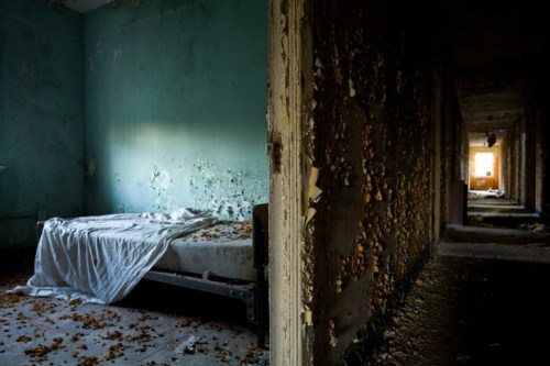 Abandoned room with a disheveled bed, peeling walls, and debris scattered on the floor, illuminated by soft light.