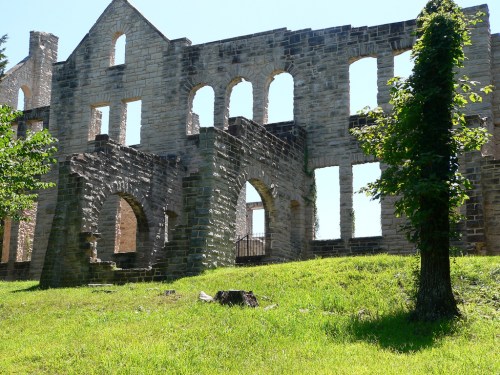 Ruins of a stone building with arched windows, surrounded by green grass and trees under a clear blue sky.