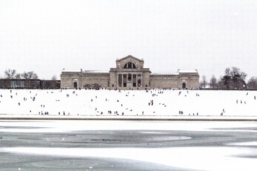 A snowy landscape with a large building in the background and people playing in the snow.