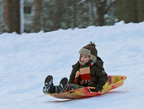 A child in winter clothing sleds down a snowy hill, wearing a hat and scarf, with a joyful expression.