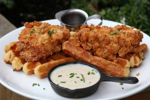 Plate of crispy fried chicken on waffles, served with a side of creamy sauce and a small cup of syrup.