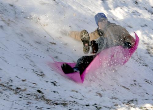 A person sledding down a snowy hill on a pink sled, captured in motion with snow flying around.