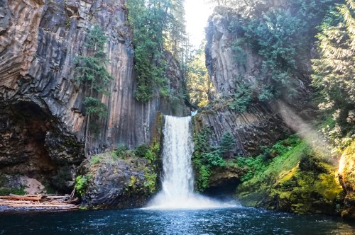 A serene waterfall cascades into a clear pool, surrounded by towering cliffs and lush greenery. Sunlight filters through trees.