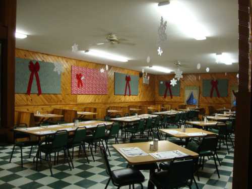 A festive dining hall with green chairs, checkered floor, and holiday decorations like bows and snowflakes on the walls.