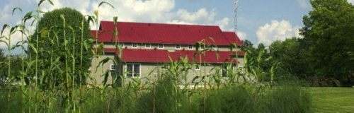 A two-story building with a red roof surrounded by tall grass and trees under a partly cloudy sky.