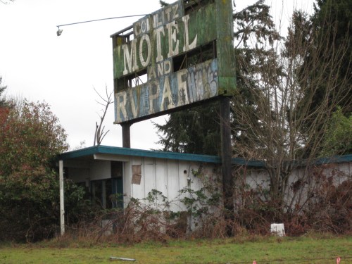 Abandoned motel with a weathered sign reading "Motel and RV Park," surrounded by overgrown vegetation.