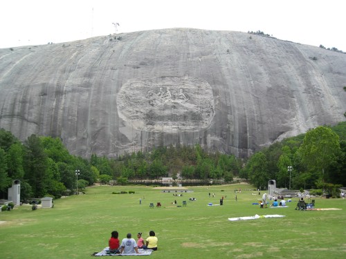 A large granite mountain with a carved relief, surrounded by green grass and people enjoying a park.