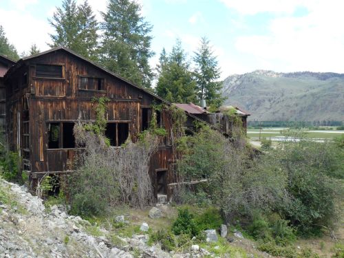 Abandoned wooden building overgrown with vegetation, set against a backdrop of mountains and a clear sky.