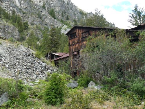 Abandoned wooden building surrounded by rocky terrain and lush greenery, set against a mountainous backdrop.