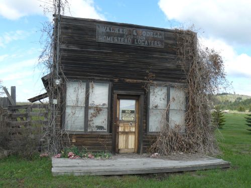 Abandoned wooden building with a sign, overgrown with vines, surrounded by grass and a wooden fence.