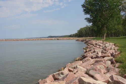 A serene lakeside view with a rocky shoreline and green trees under a partly cloudy sky.