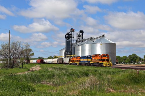 A colorful train passes by large grain silos under a blue sky with fluffy clouds, surrounded by green grass.