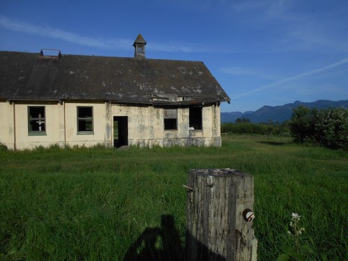 An old, weathered building stands in a grassy field under a clear blue sky, surrounded by mountains.