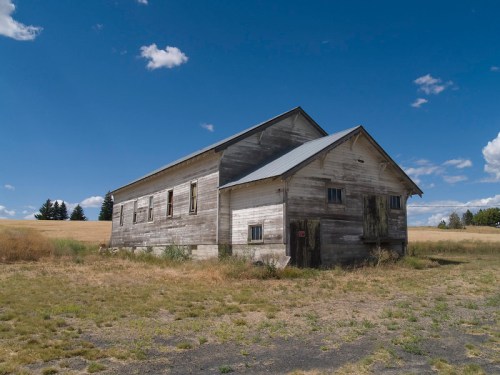 An old, weathered wooden house stands in a grassy field under a blue sky with scattered clouds.