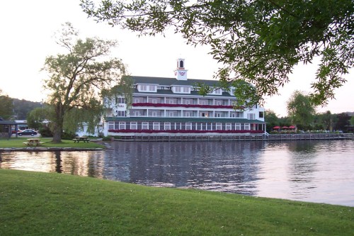 A charming lakeside hotel with a green and white facade, surrounded by trees and reflecting in the calm water.