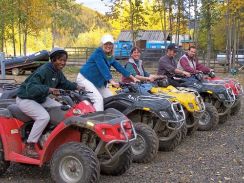 Five people sit on ATVs in a scenic outdoor setting with trees and a building in the background.