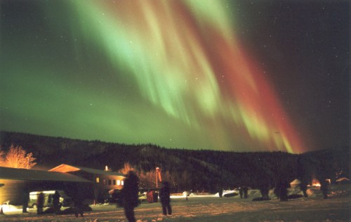 A vibrant aurora borealis lights up the night sky over a snowy landscape, with silhouettes of people in the foreground.