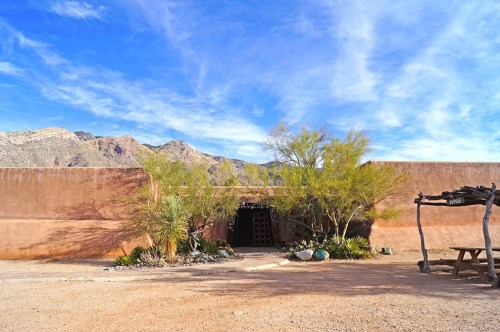 A desert landscape featuring a rustic adobe wall, entrance, and surrounding greenery with mountains in the background.