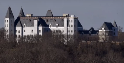 A large, unfinished castle-like structure surrounded by trees against a clear sky.