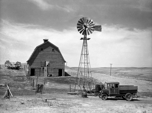 A vintage black-and-white photo of a barn, windmill, and an old truck on a rural landscape.