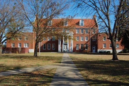 A brick building with a central entrance, surrounded by trees and a pathway, on a clear day with autumn foliage.