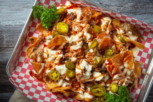 A tray of loaded nachos topped with pulled pork, jalapeños, drizzled with sauce and sour cream, garnished with parsley.