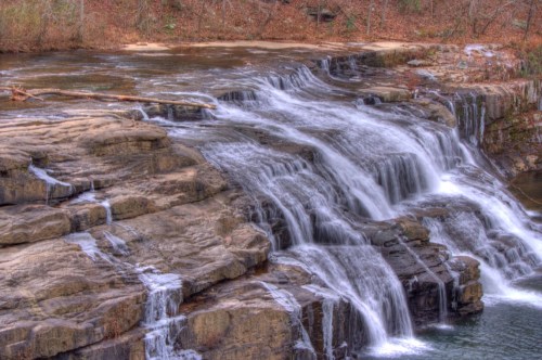 A serene waterfall cascading over rocky ledges, surrounded by trees and a calm river.