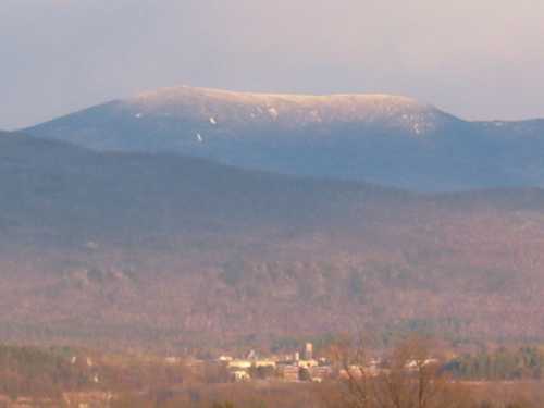 A distant mountain peak with a light dusting of snow, overlooking a valley with trees and a small town below.