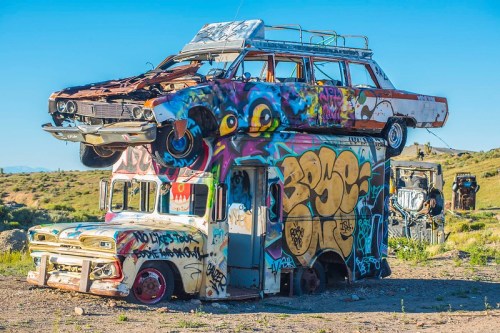 A colorful stack of graffiti-covered cars, with one car on top of a painted truck in a desert landscape.