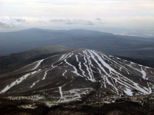 Aerial view of a snow-covered mountain with ski trails winding down its slopes, surrounded by distant hills and clouds.