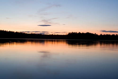 Serene lake at sunset, reflecting soft hues of orange and blue with silhouettes of trees along the horizon.