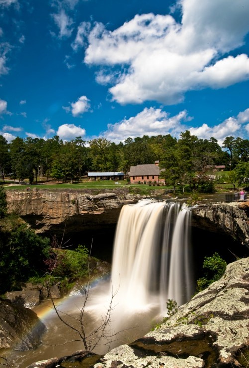 A waterfall cascades over rocky cliffs, surrounded by lush greenery and a bright blue sky with fluffy clouds.