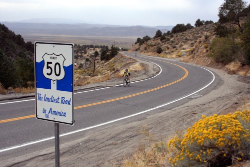 A cyclist rides along a winding road with a sign reading "Hwy 50: The Loneliest Road in America" in a desert landscape.