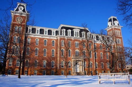 Historic brick building with clock towers, surrounded by snow-covered trees and a white bench in a winter landscape.