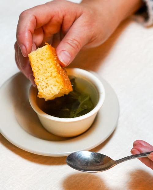 A hand holds a piece of cornbread above a small bowl of greens, with a spoon resting nearby on a table.