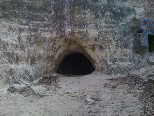 A dark cave entrance in a rocky, sandy landscape, surrounded by weathered stone and faint graffiti.