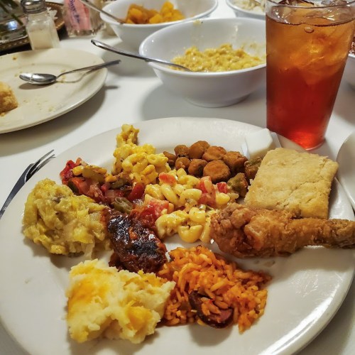 A plate of Southern comfort food featuring fried chicken, macaroni, rice, okra, and cornbread, with iced tea in the background.