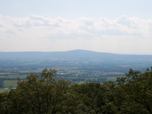 A panoramic view of rolling hills and valleys under a cloudy sky, with trees in the foreground.
