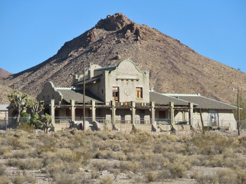 Abandoned stone building in a desert landscape, with a mountain in the background and sparse vegetation around.