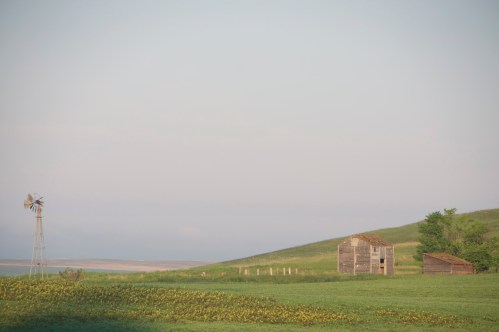 A serene landscape featuring a windmill, a weathered barn, and rolling green hills under a soft sky.