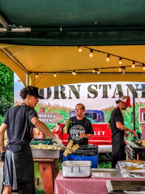 A corn stand with three workers preparing food under a tent, with string lights and a vintage truck in the background.