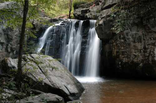 A serene waterfall cascades over rocks into a calm pool, surrounded by lush greenery and rugged terrain.