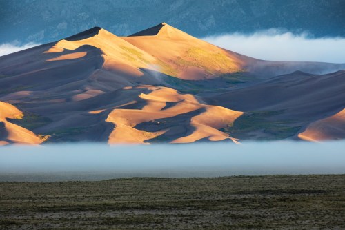 Golden sand dunes rise above a layer of mist, set against a backdrop of mountains and a clear blue sky.