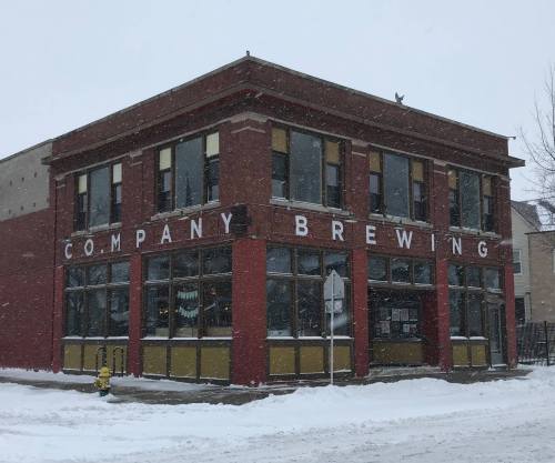 A red brick building with large windows, featuring the sign "Company Brewing," surrounded by snow in a winter scene.