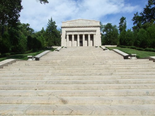 A grand stone monument with columns, set on a staircase surrounded by greenery under a blue sky.