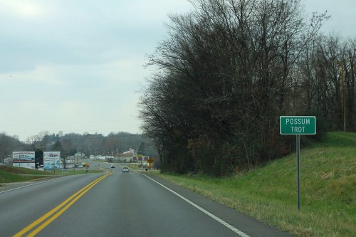 Road sign for "Possum Trot" along a two-lane highway, with trees and a small town visible in the background.